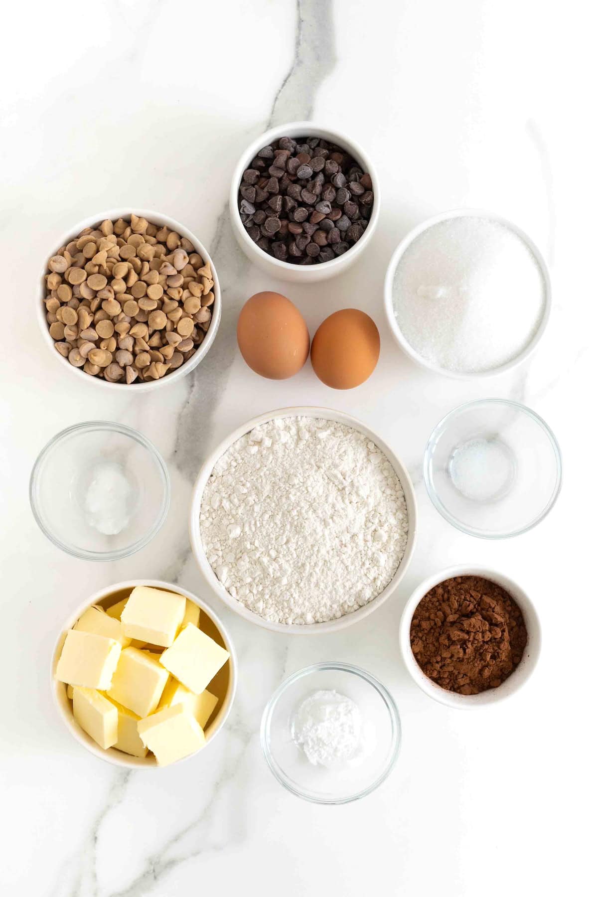 Ingredients to make chocolate peanut butter chip cookies in small glass containers on a white marble counter.