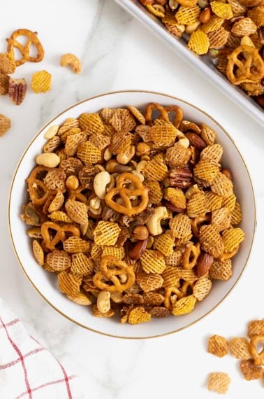 A large white mixing bowl filled with a snack mix made of cereals, nuts and pretzels.