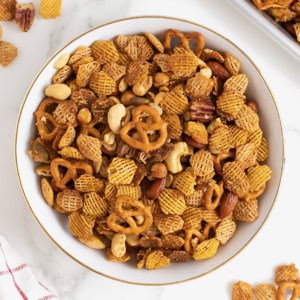 A large white mixing bowl filled with a snack mix made of cereals, nuts and pretzels.