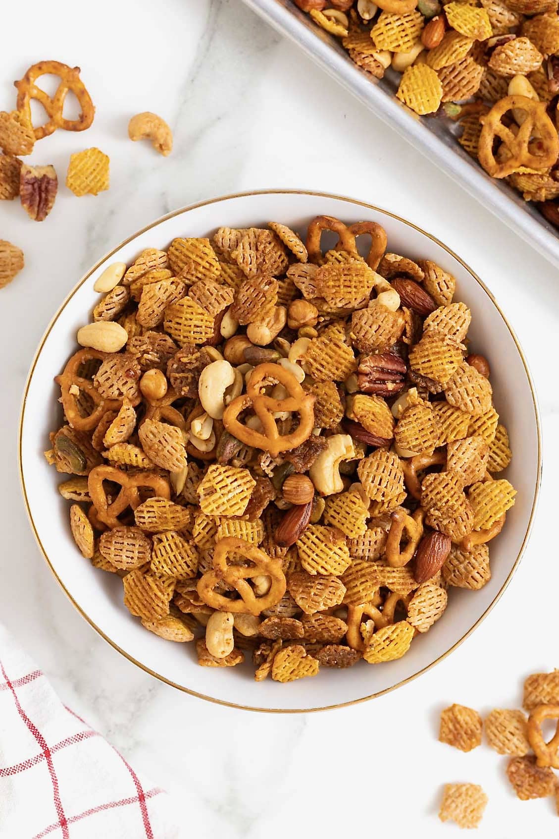 A large white mixing bowl filled with a snack mix made of cereals, nuts and pretzels.