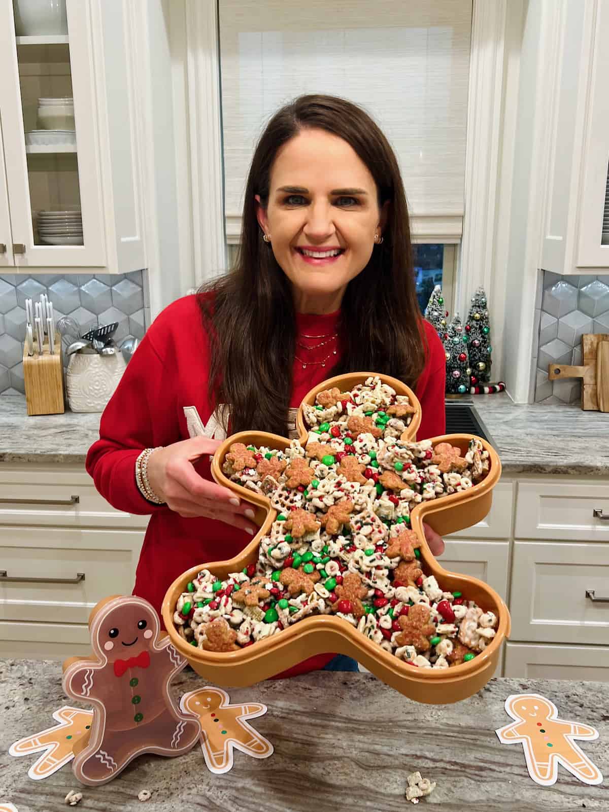 Maegan Brown wearing a red sweater holding a large gingerbread man shaped dish filled with snack mix.