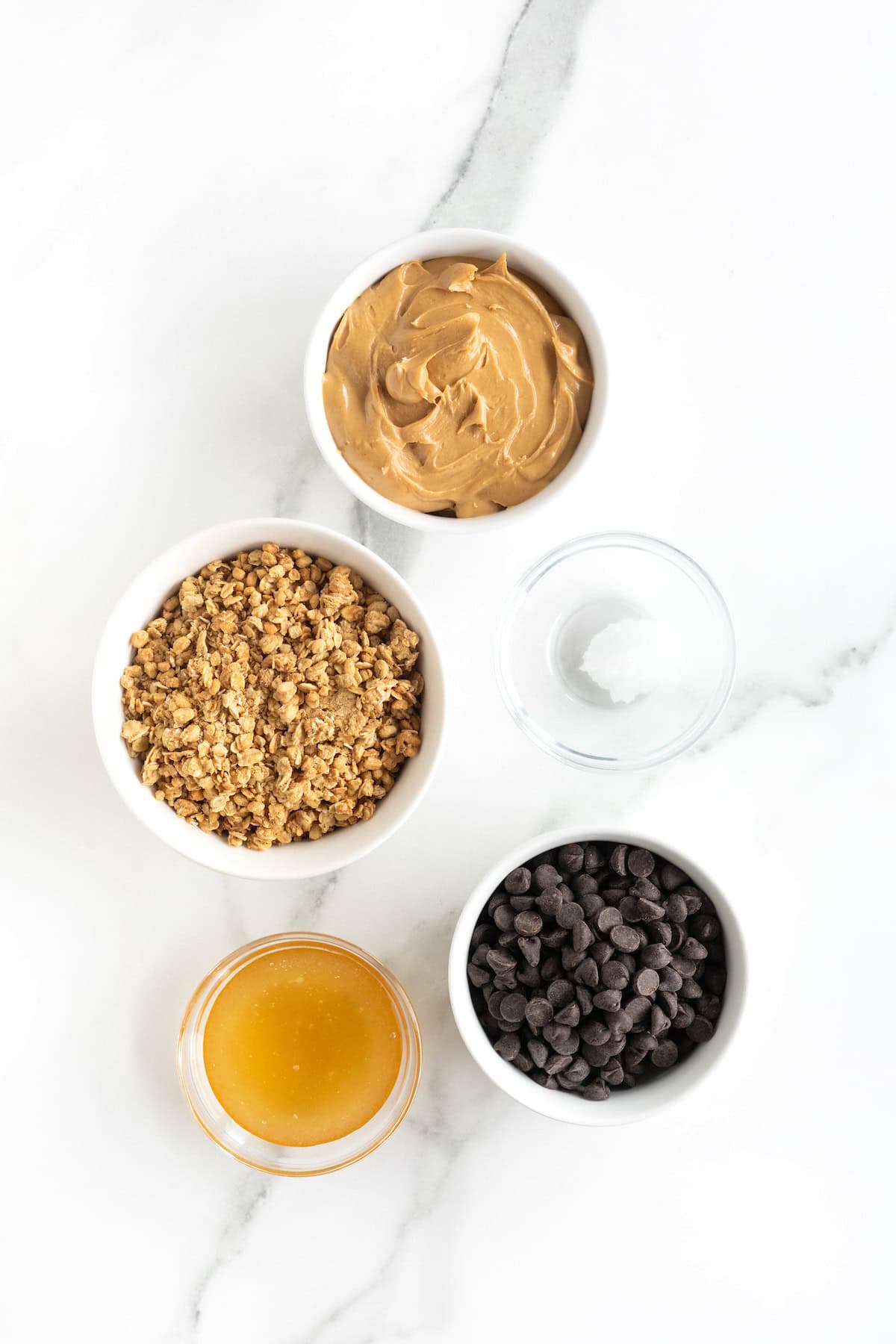 Ingredients to make healthy peanut butter balls in small glass dishes on a white marble counter.