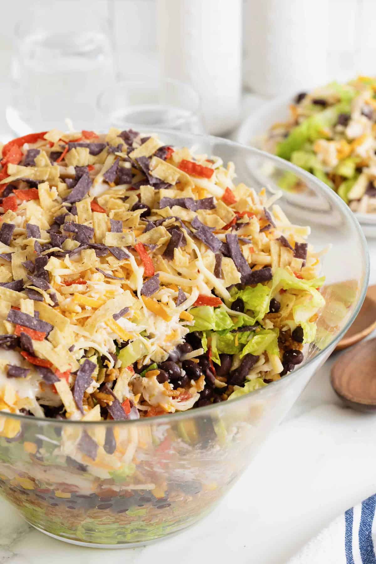 A layered chicken taco salad in a large glass dish showing layers of salad, beans, corn, sour cream, avocado, chicken, cheese and tortilla strips.