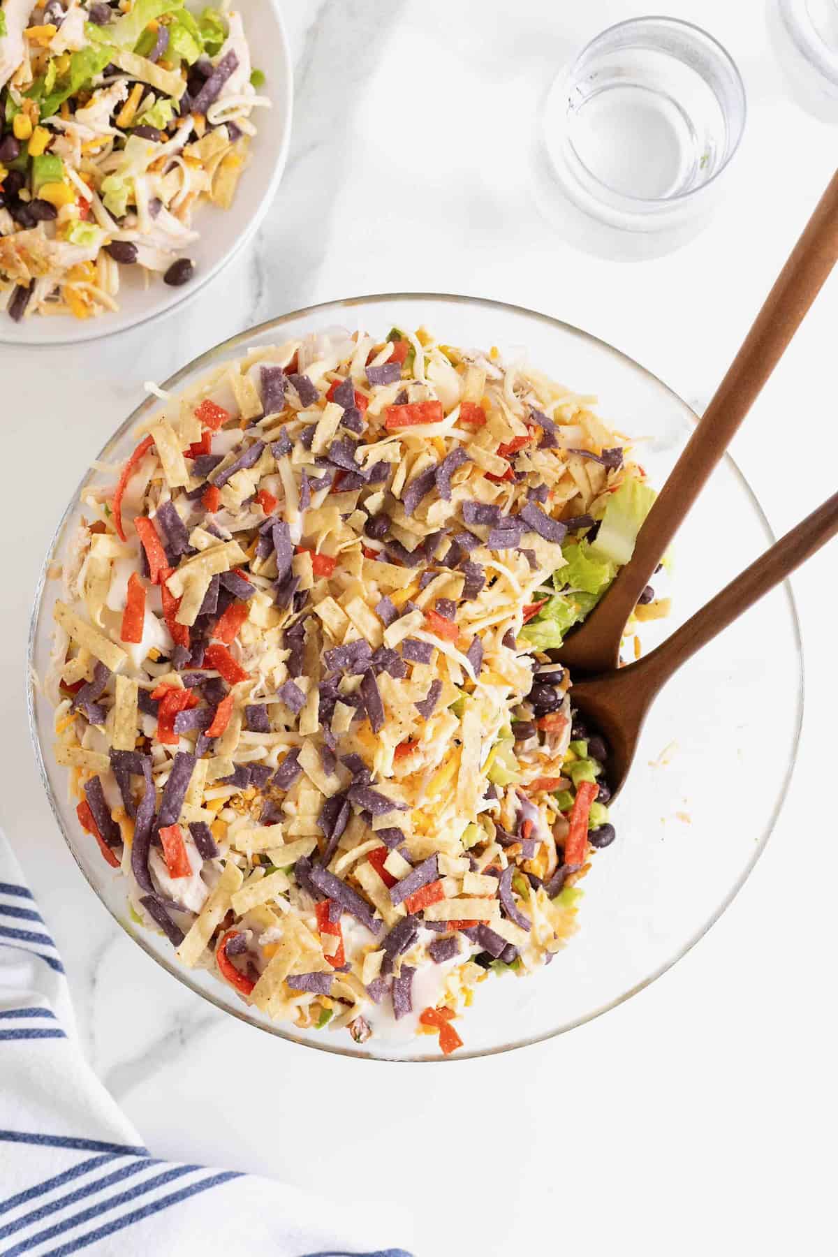 A layered chicken taco salad in a large glass dish showing layers of salad, beans, corn, sour cream, avocado, chicken, cheese and tortilla strips.