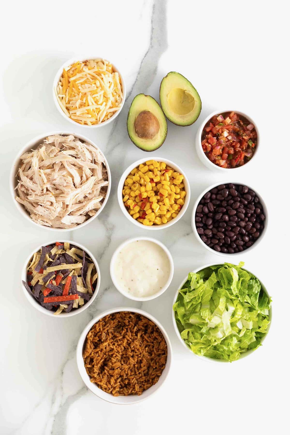 Ingredients to make layered chicken taco salad in small white dishes on a white marble counter.