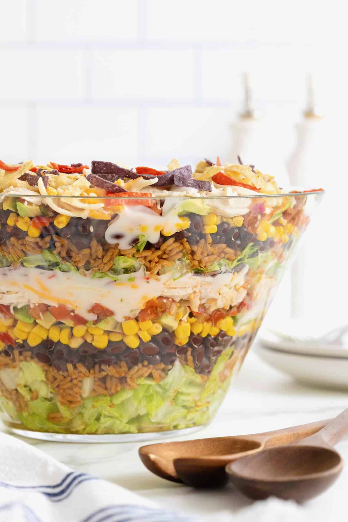 A layered chicken taco salad in a large glass dish showing layers of salad, rice, beans, corn, avocado, tomato, sour cream, avocado, chicken, cheese and tortilla strips.