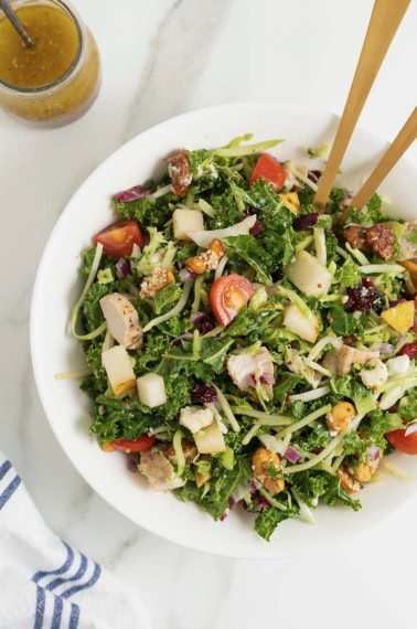 A large white bowl of green salad with cut up tomatoes, shredded cheese and chunks of chicken.
