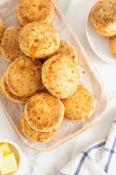A pile of cheddar biscuits on non oblong cutting board covered in parchment.