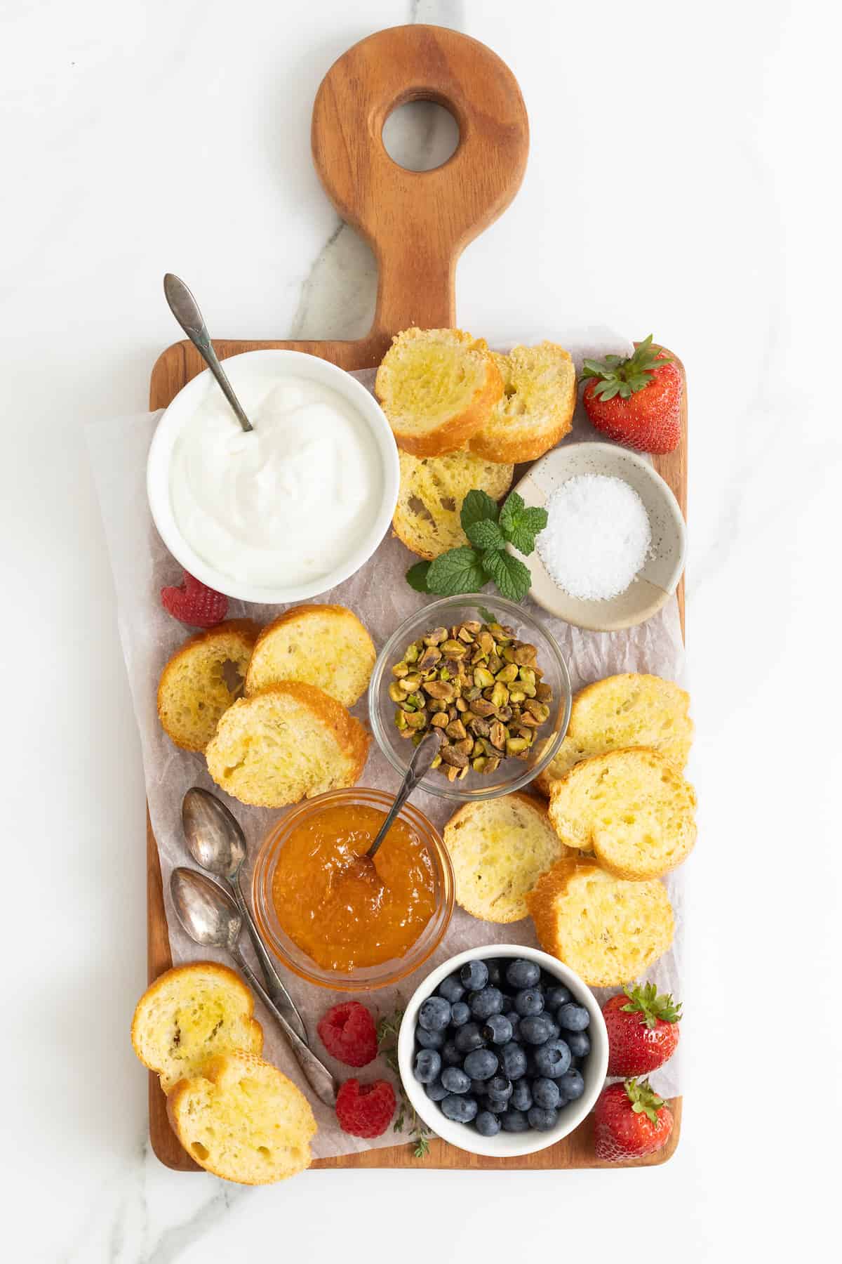 A rectangular food board with a white dish of labneh in the top left corner, a dish of pistachios in the middle, and jams, toppings and toast surrounding.