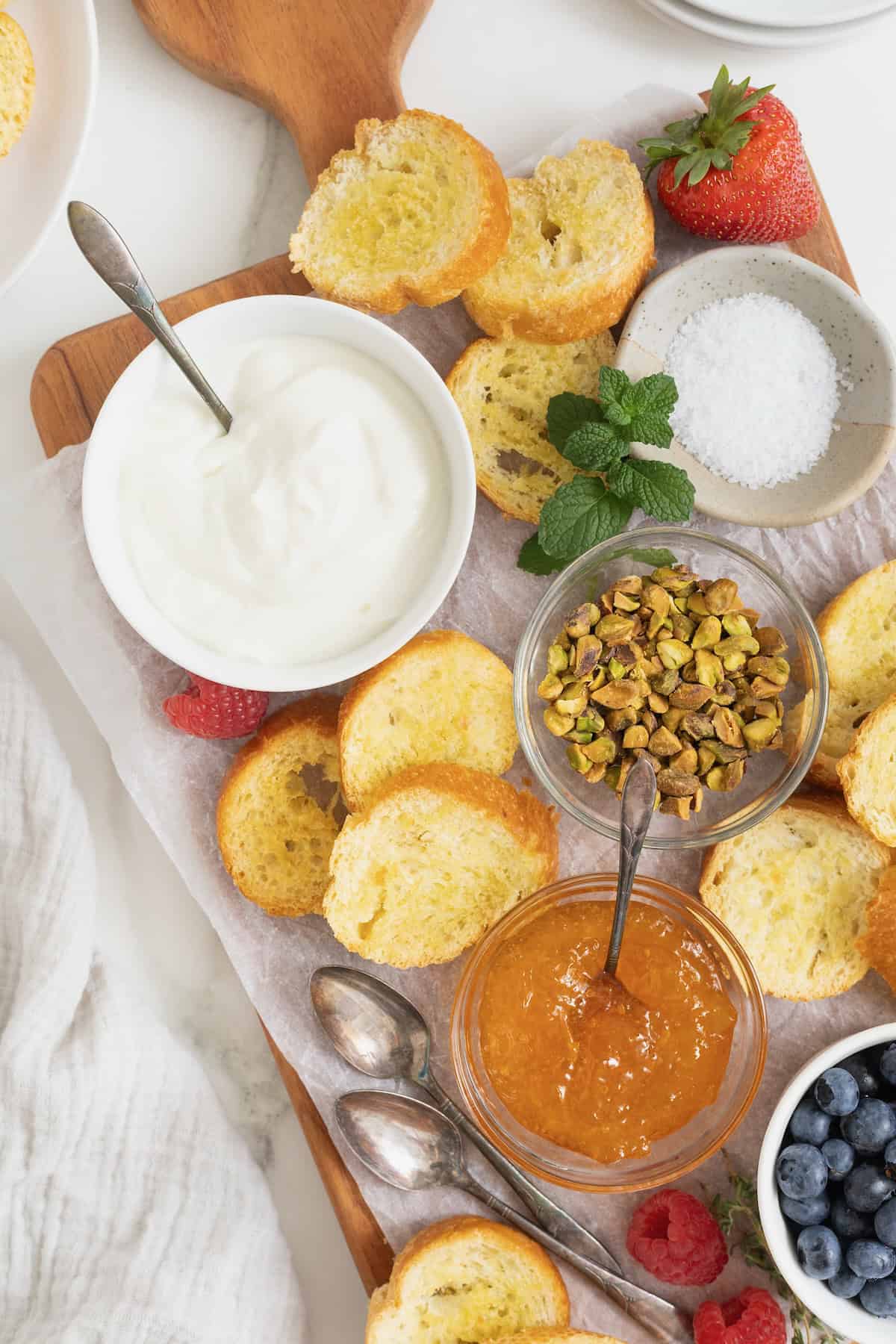 A rectangular food board with a white dish of labneh in the top left corner, a dish of pistachios in the middle, and jams, toppings and toast surrounding.