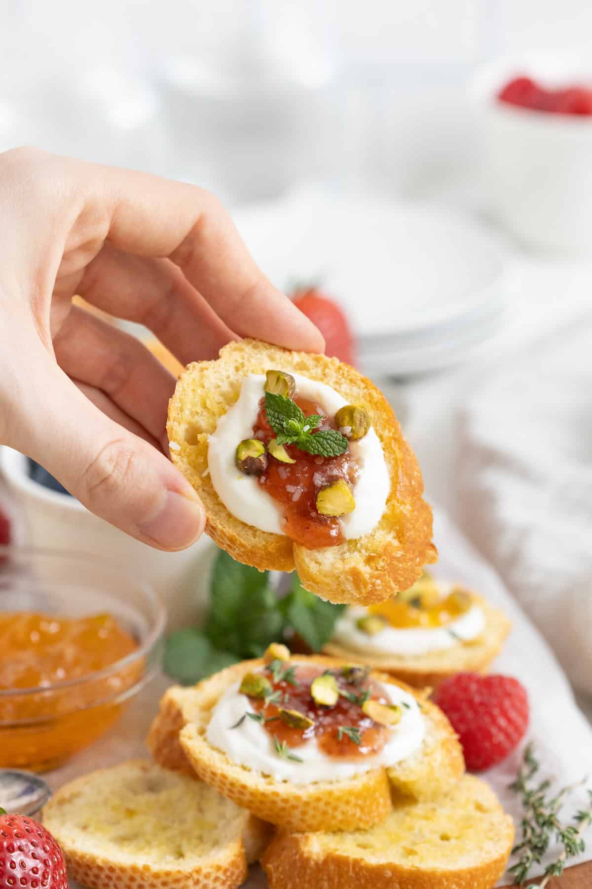A hand holding a piece of crostini topped with a dollop of labneh, jam, and a few chopped pistachios.