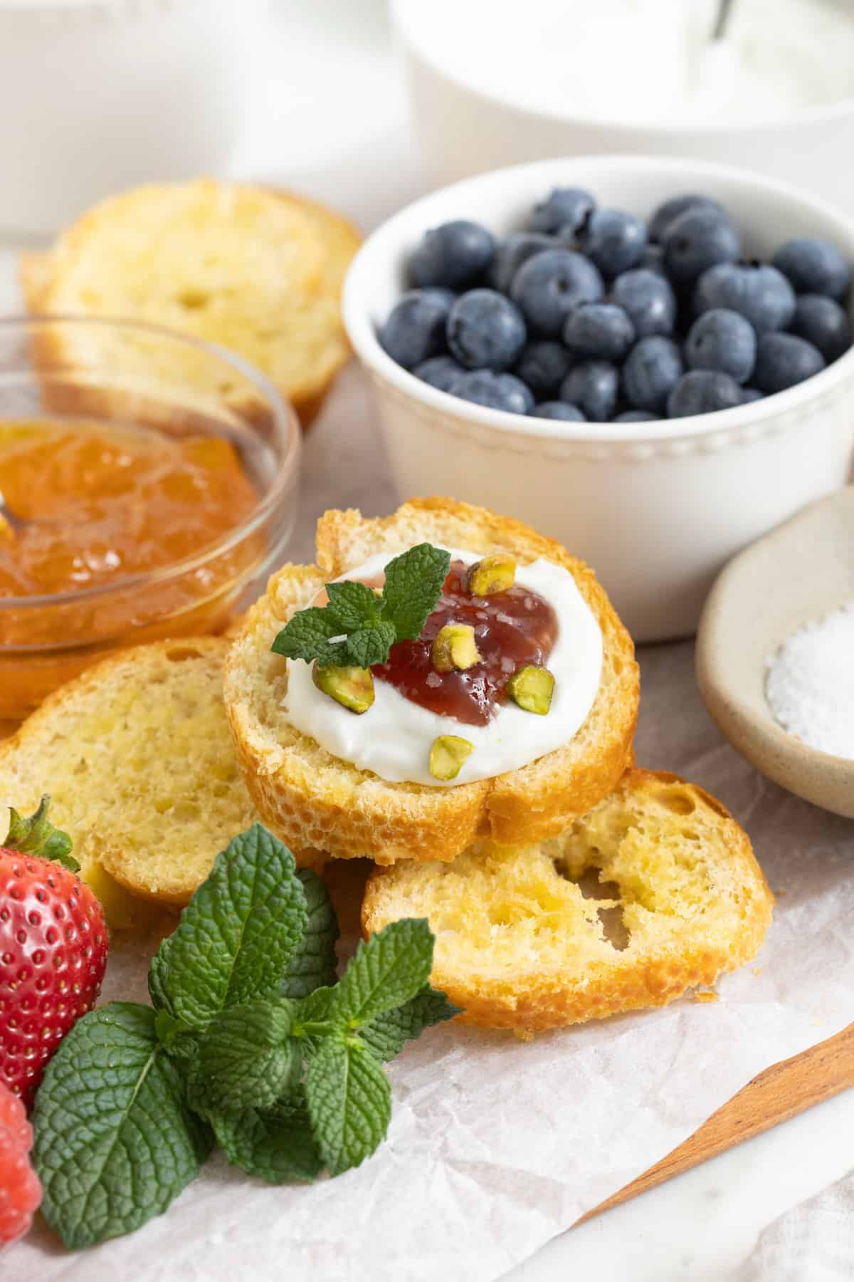 A piece of crostini topped with labneh, jam, chopped pistachios and a sprig of mint on top of a pile of baked crostini.