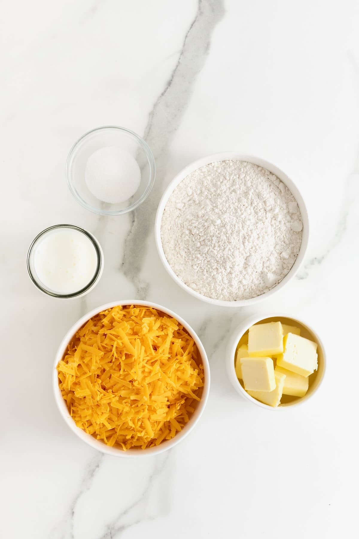 Ingredients to make flaky cheddar biscuits in small glass dishes on a white marble counter.