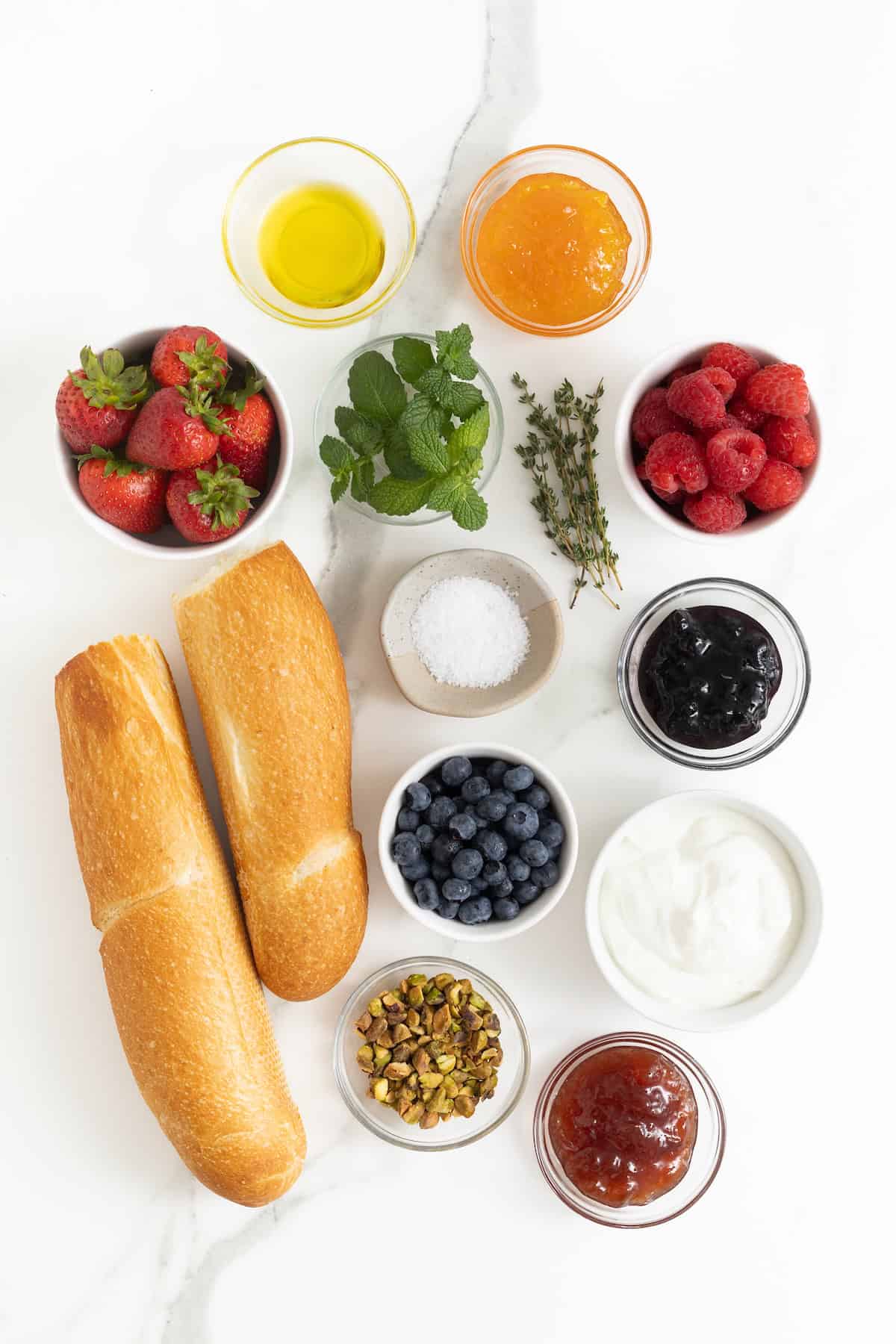 A long baguette cut in half next to the ingredients to make a labneh toast board in small glass dishes on a white marble counter.