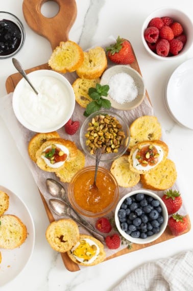 A rectangular food board with a white dish of labneh in the top left corner, a dish of pistachios in the middle, and jams, toppings and toast surrounding.