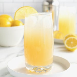 A tall glass of lemonade cocktail on a rimmed white tray on a white marble counter. In the background is a white bowl filled with lemons.