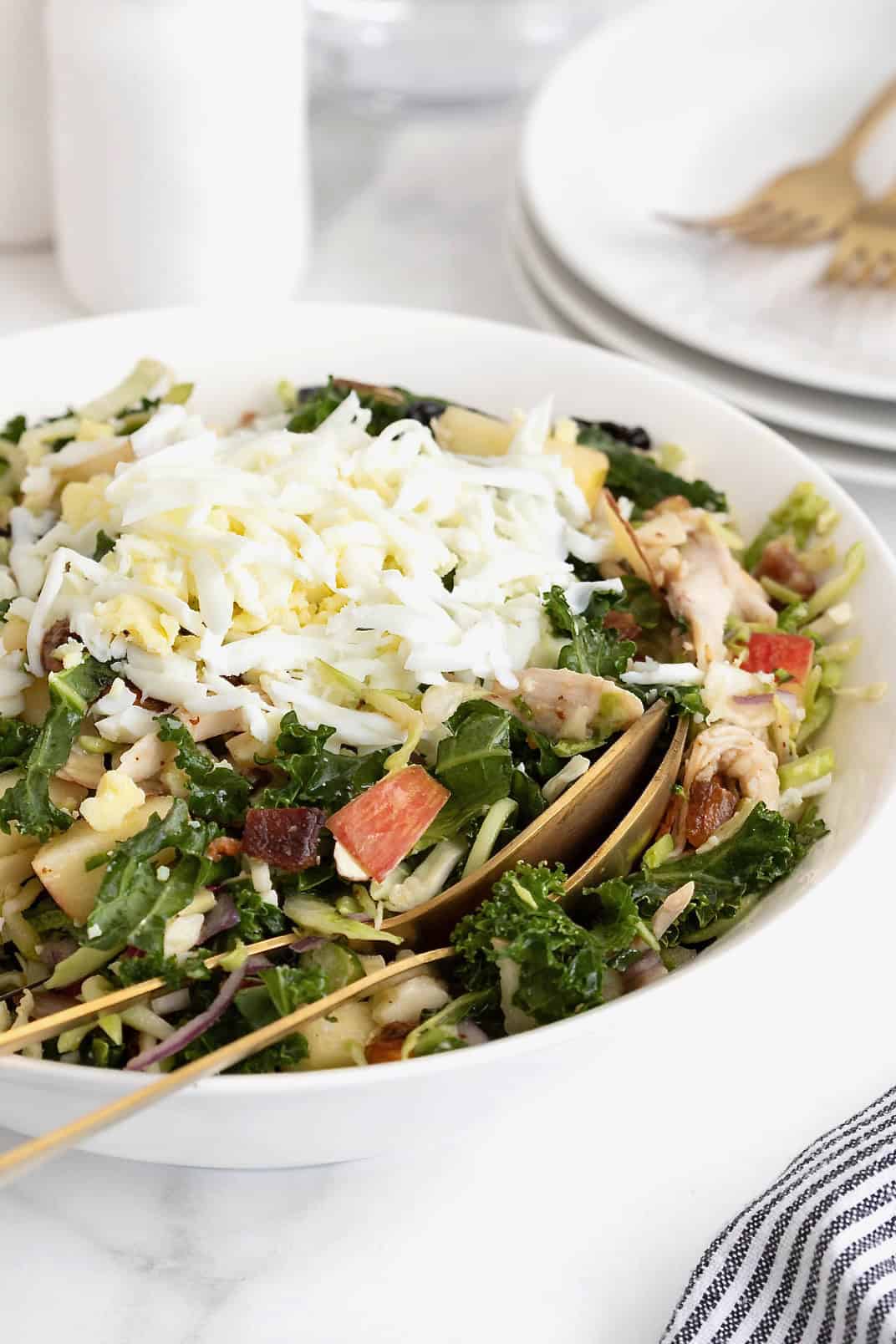A large white serving bowl of kale salad with two gold toned spoons inside.