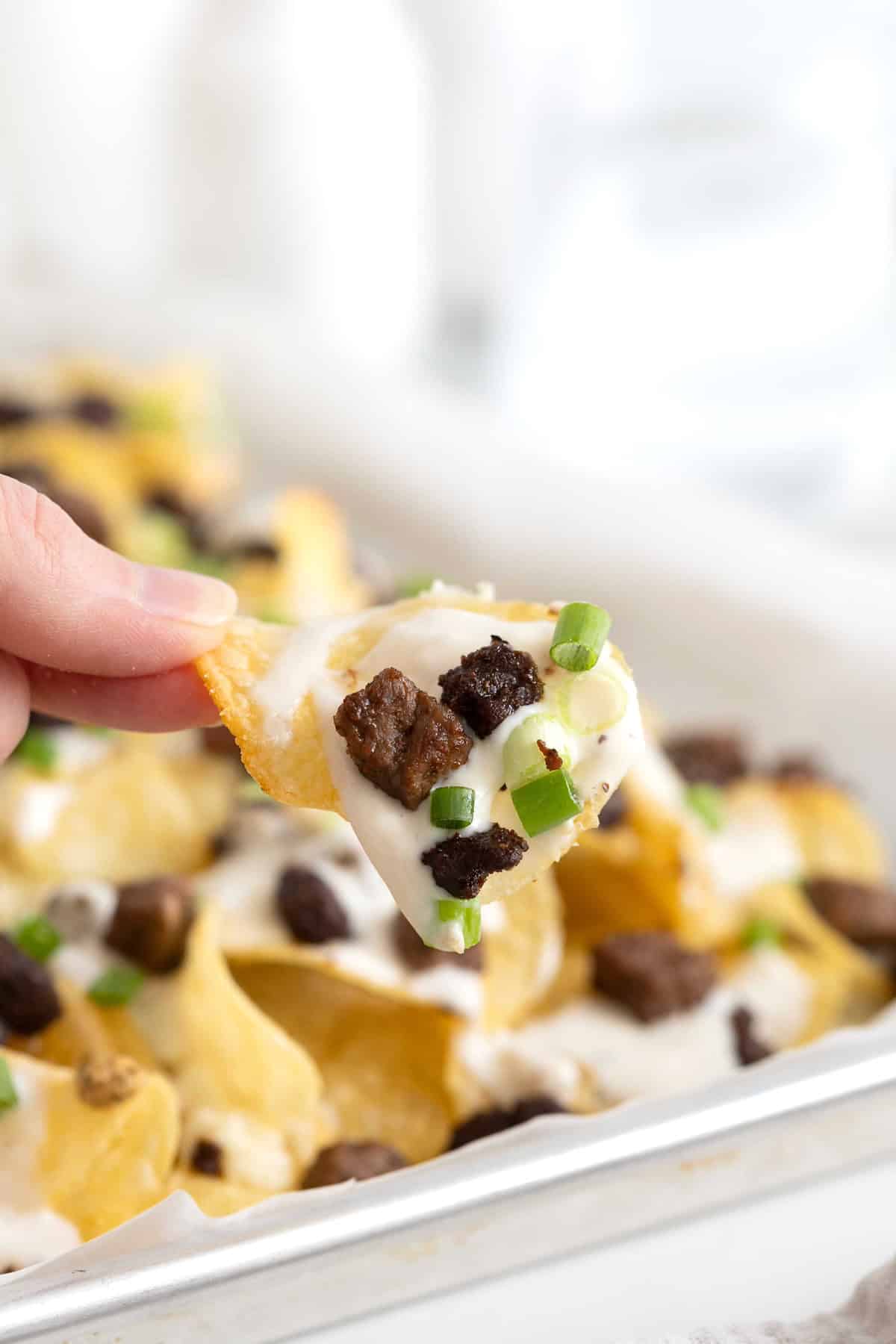 A hand holding a potato chip with steak and blue cheese on it between two fingers.