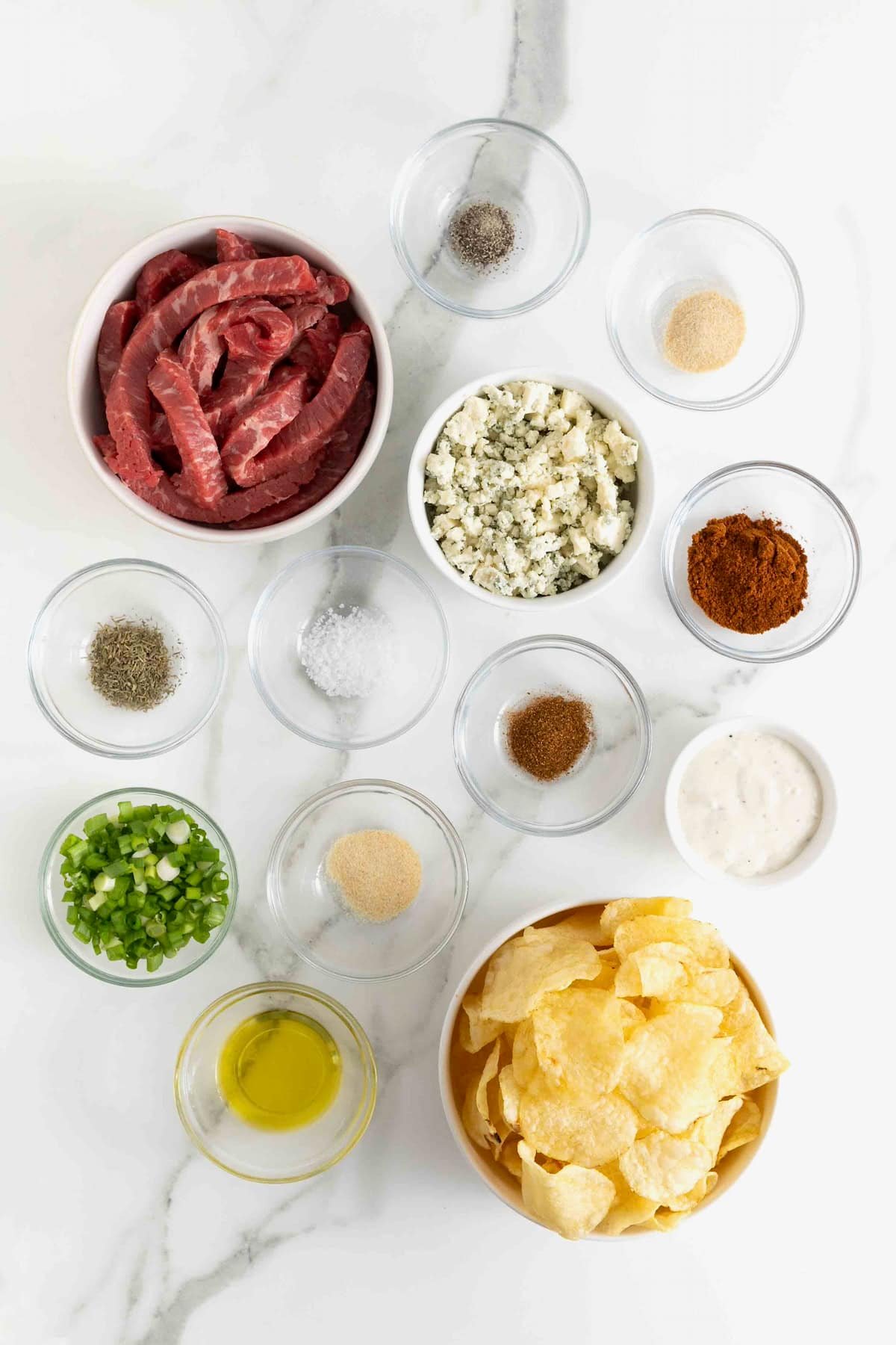 Ingredients to make steak and blue cheese nachos in small glass dishes on a white marble counter.