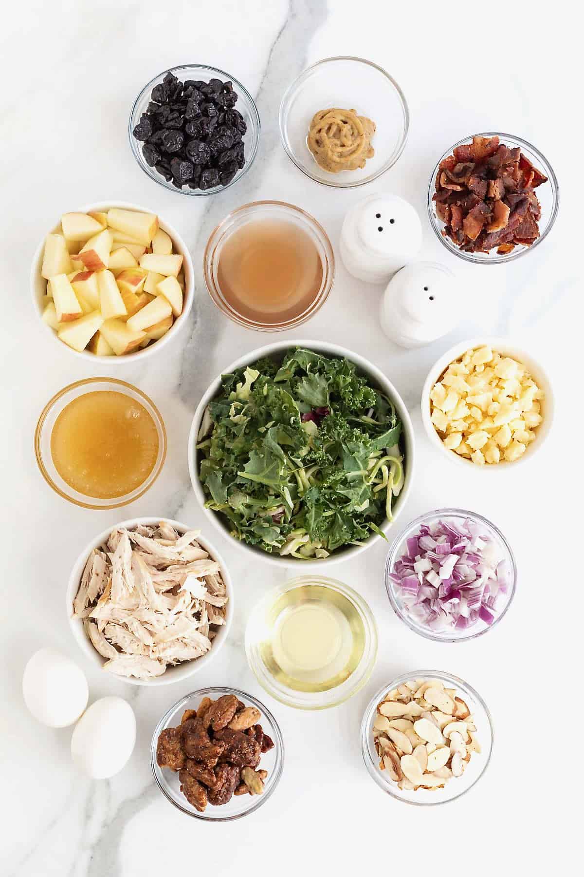 Ingredients to make a kale and roasted chicken salad in small glass dishes on a white marble counter.