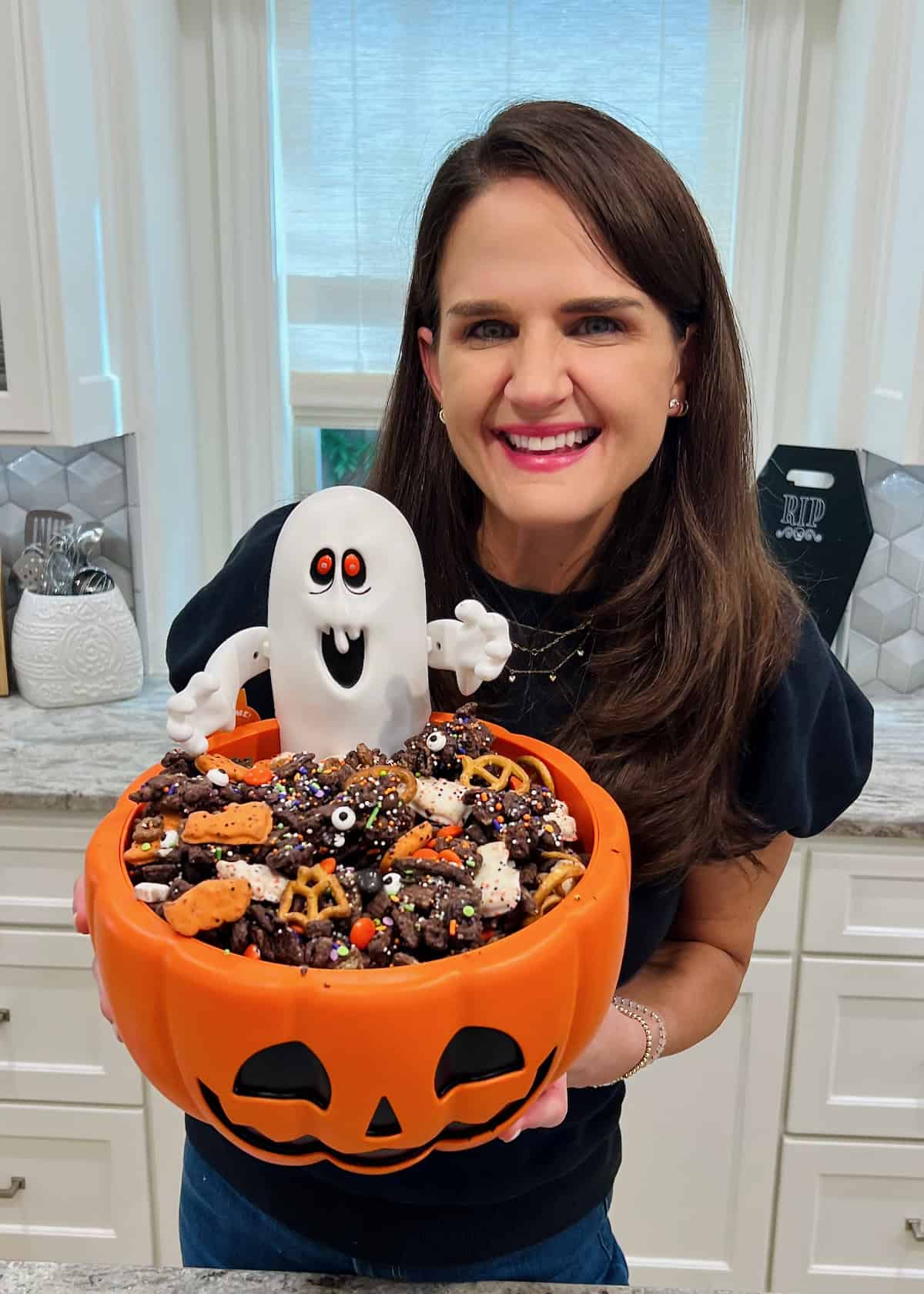 Maegan Brown wearing a black short sleeve top holding a Jack O' Lantern dish of Halloween Snack Mix.