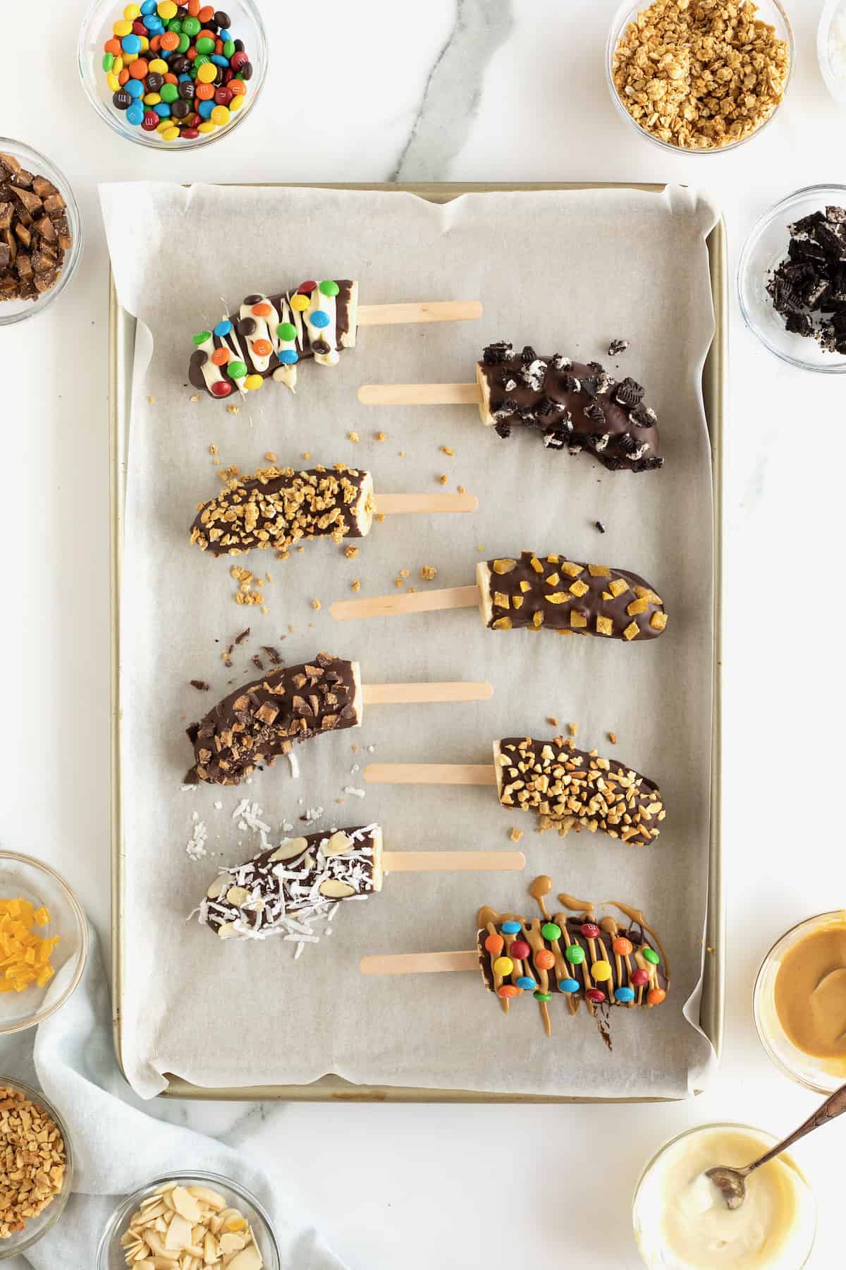 Frozen bananas with popsicles sticks covered in chocolate and mini M&Ms, mini chocolate chips, crushed peanuts and coconut.