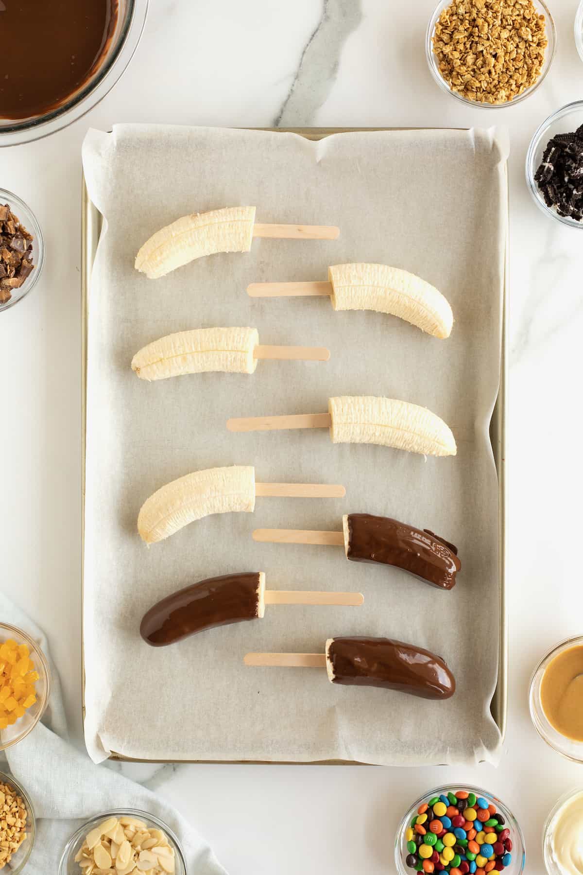 Bananas on popsicles sticks covered in melted chocolate on a parchment lined baking tray.