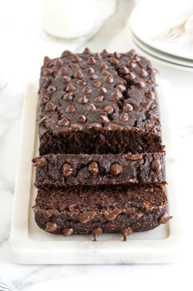 A loaf of chocolate banana bread studded with chocolate chips on top. The loaf is sitting on a white cutting board on a white marble counter.