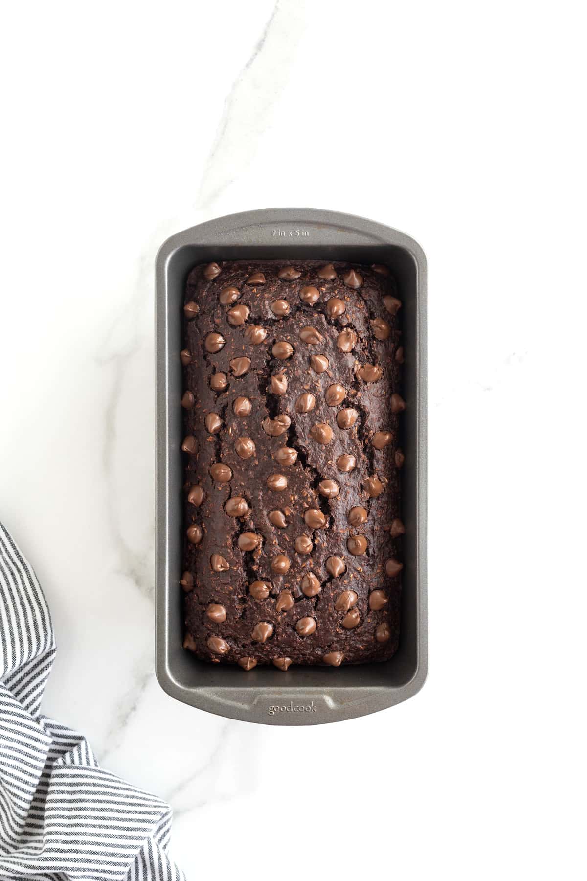 A baked loaf of chocolate banana bread studded with chocolate chips in a dark metal loaf pan on a white marble counter.
