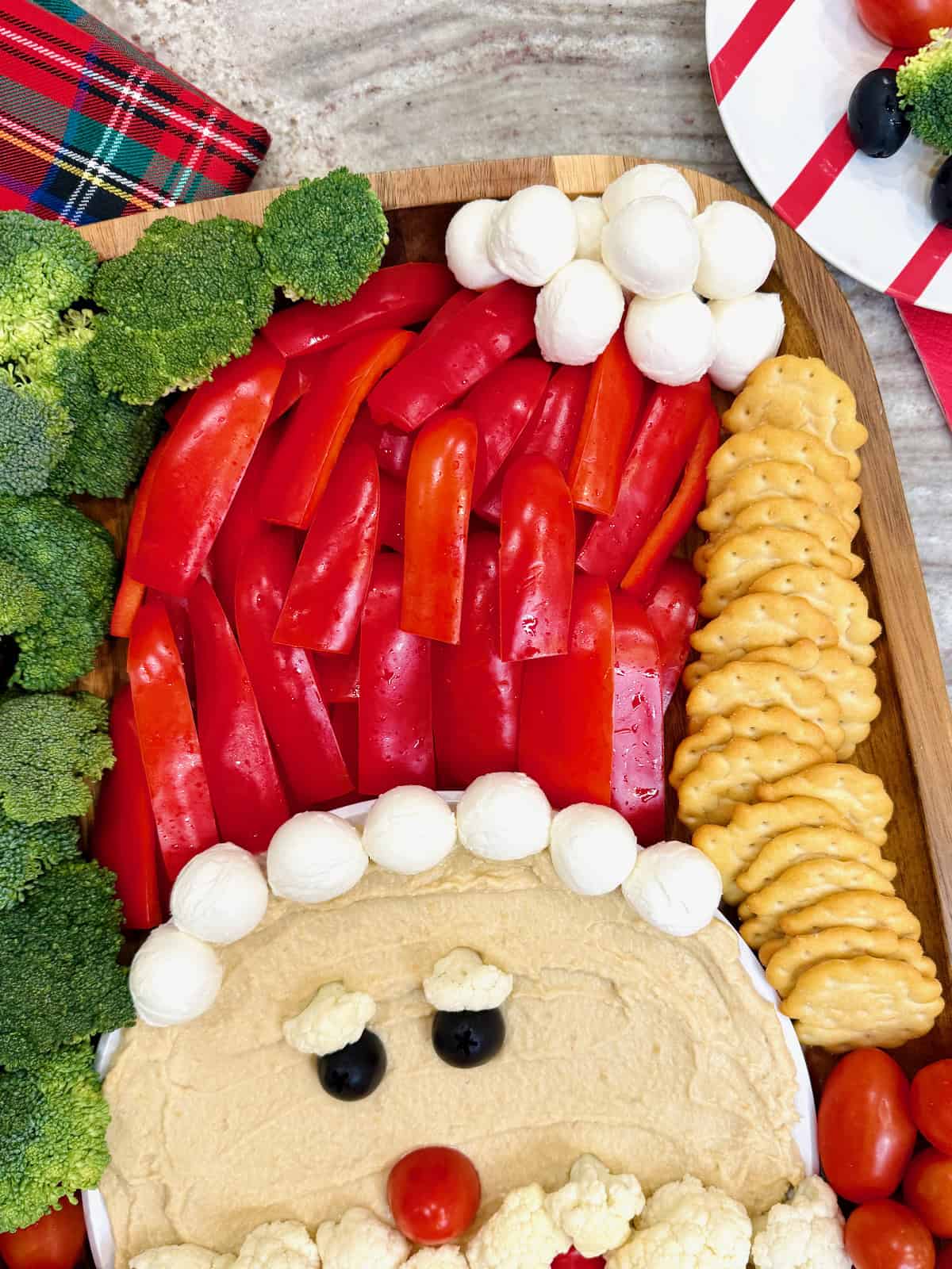 A snack board with Hummus, cauliflower, cherry tomatoes, broccoli, and crackers forming the shape of Santa's face.