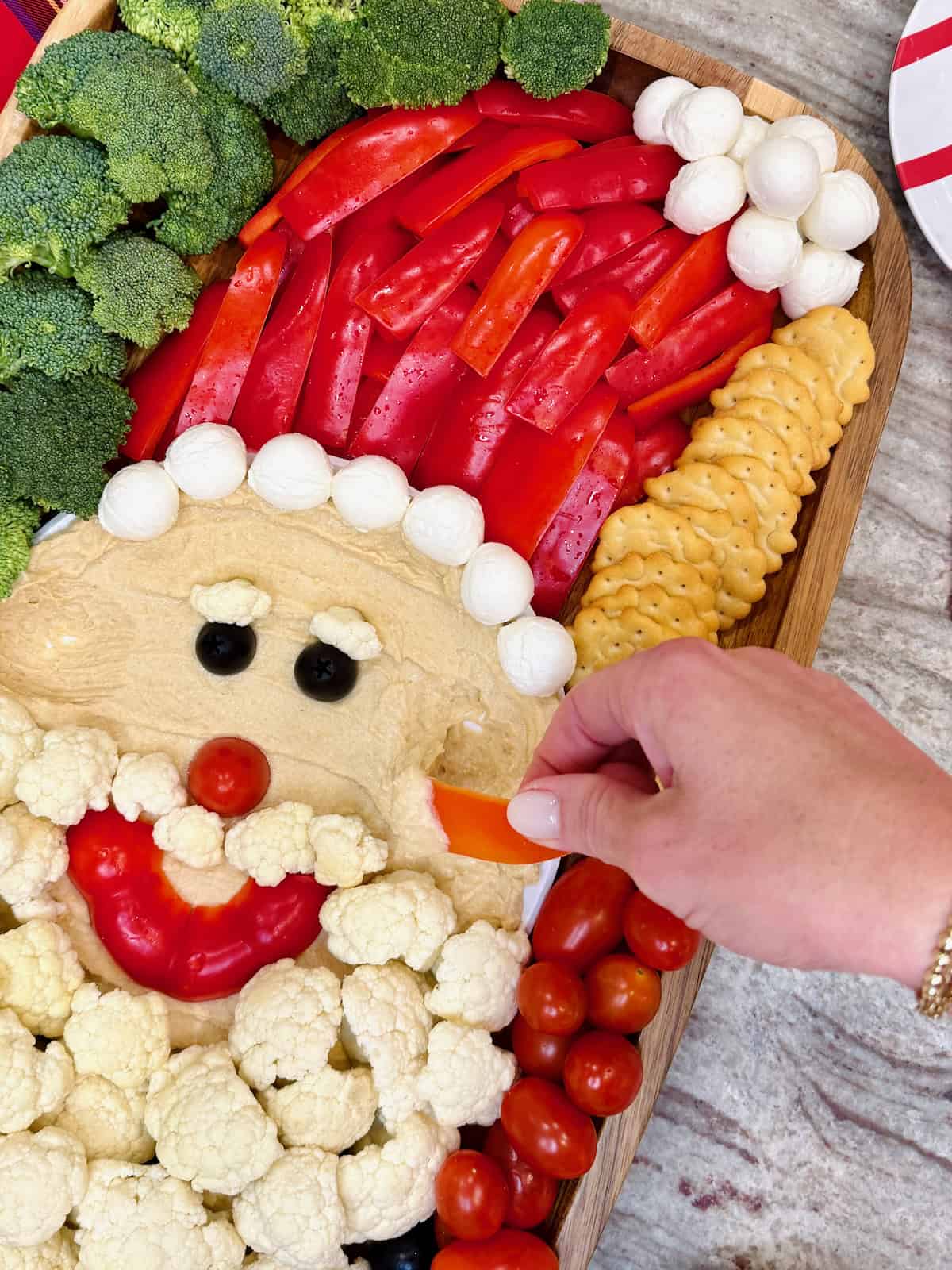 A hand dipping a red pepper strip into a bowl of hummus on a Santa Snack Board.