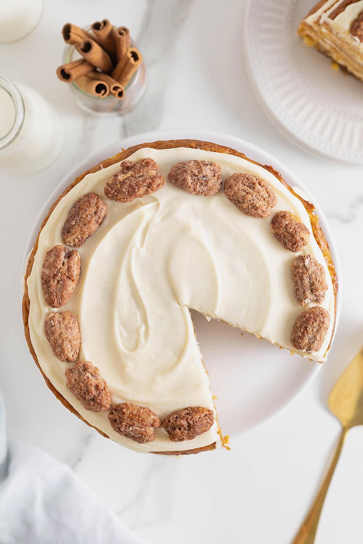 A cake with white buttercream frosting and candied pecans as seen from the top. A piece is cut out from the cake.