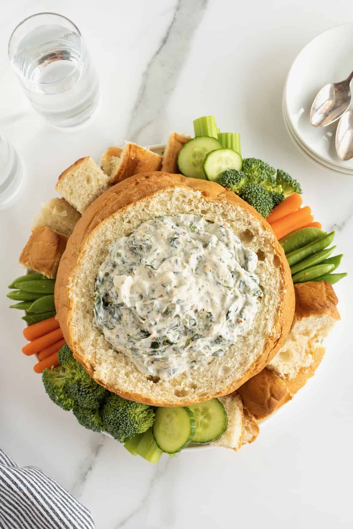 Creamy spinach in a bread bowl surrounded by fresh vegetables and bread chunks.
