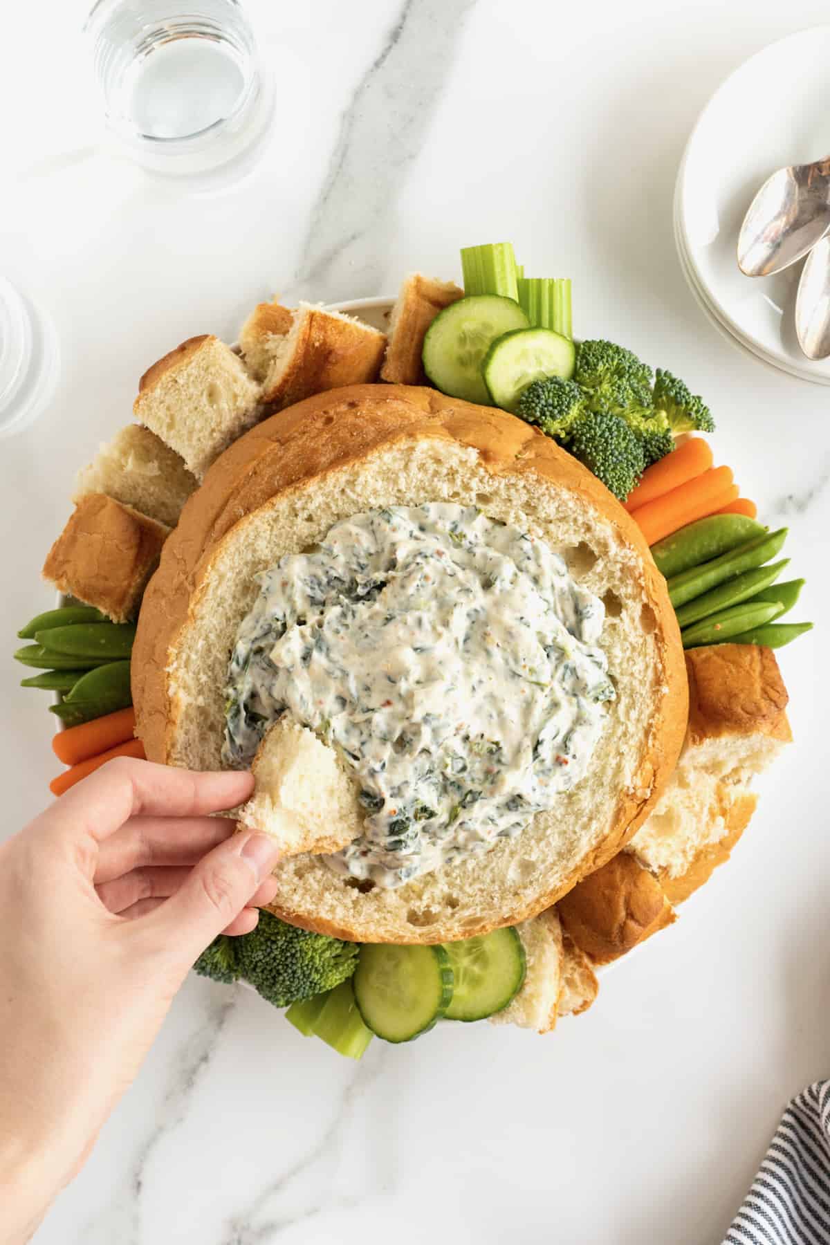 Creamy spinach dip in a bread bowl surrounded by fresh vegetables and bread chunks.