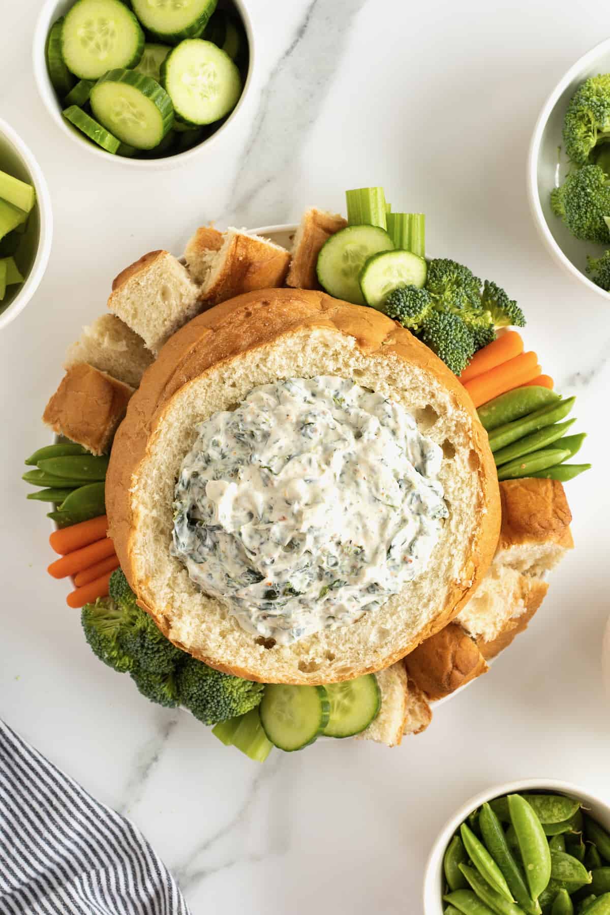Creamy spinach dip in bread bowl surrounded by fresh vegetables and bread chunks.