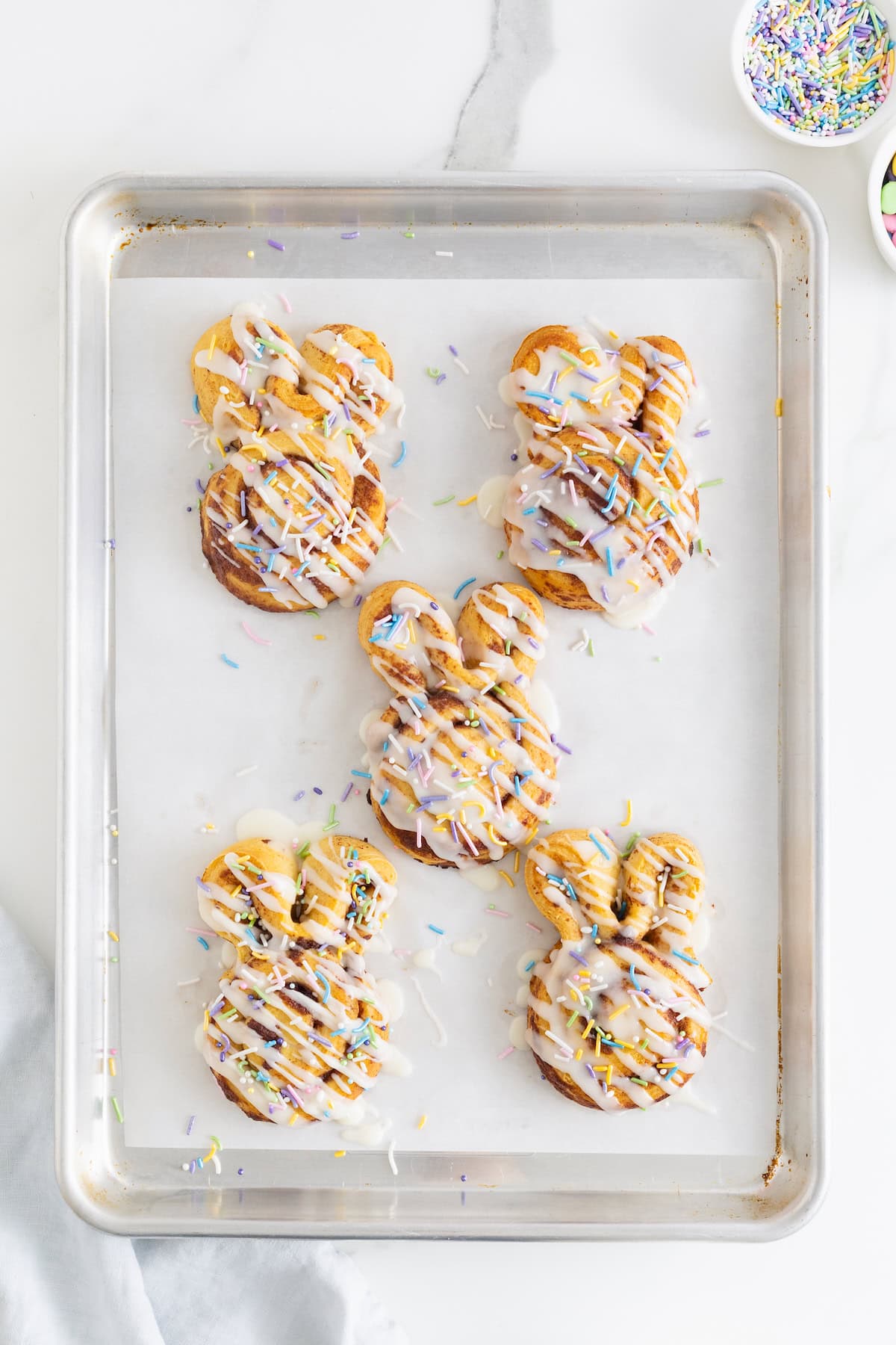 Five Easter bunny shaped cinnamon rolls decorated with white icing and pastel sprinkles.