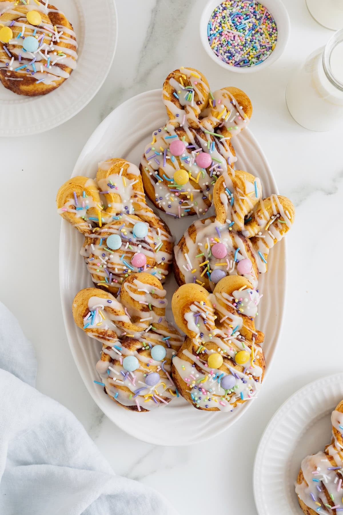 Five Easter bunny shaped cinnamon rolls decorated with white icing, pastel sprinkles and Easter M&Ms on an oblong white platter.
