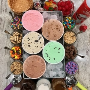 A dessert board feature ice cream sundae makings including ice cream, toppings, waffle bowls.