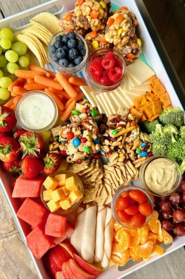 A white snack tray filled with fruits, cheeses, popcorn balls and snack bars.