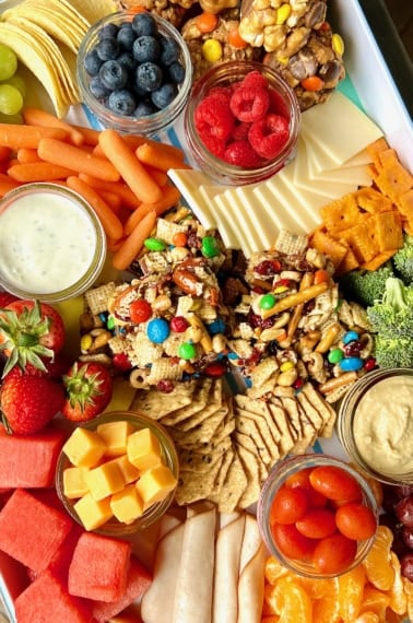 A white snack tray filled with fruits, cheeses, popcorn balls and snack bars.