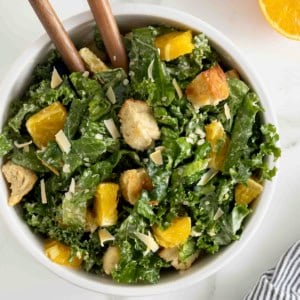 A white salad bowl of citrus caesar salad. Two wooden spoon handles are sticking out.