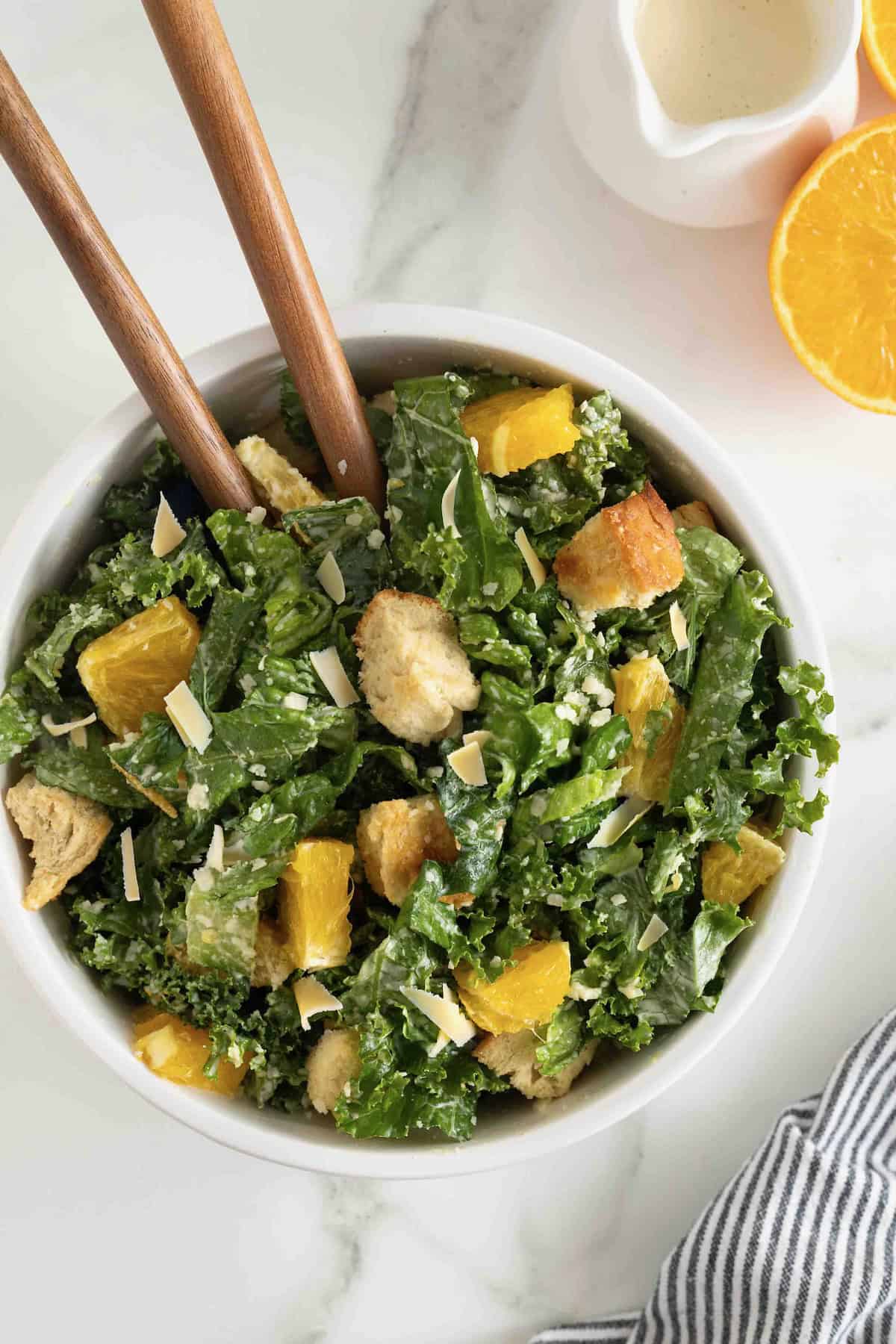 A white salad bowl of citrus caesar salad. Two wooden spoon handles are sticking out.