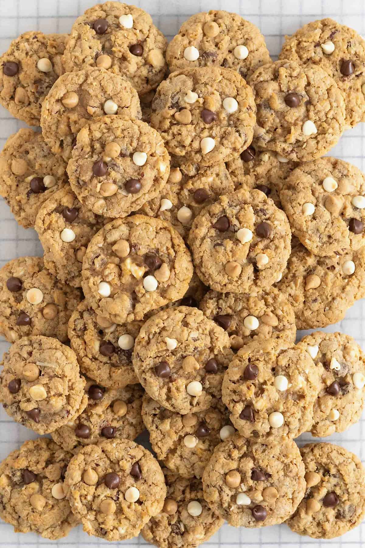 A pile of cookies on a parchment lined cooling rack. The cookies have chocolate chips, white chocolate chips, butterscotch chips and other add ins.
