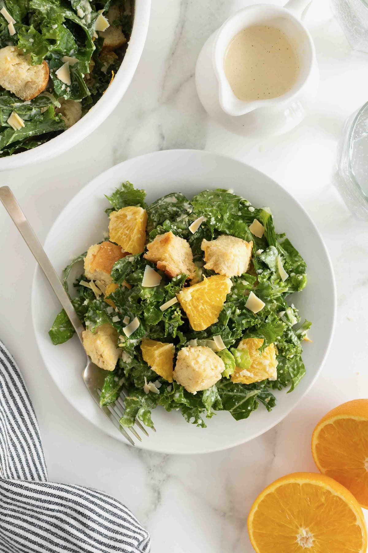 A white salad plate heaped with. citrus caesar salad. There is a fork resting on the plate and a halved orange to the bottom right.