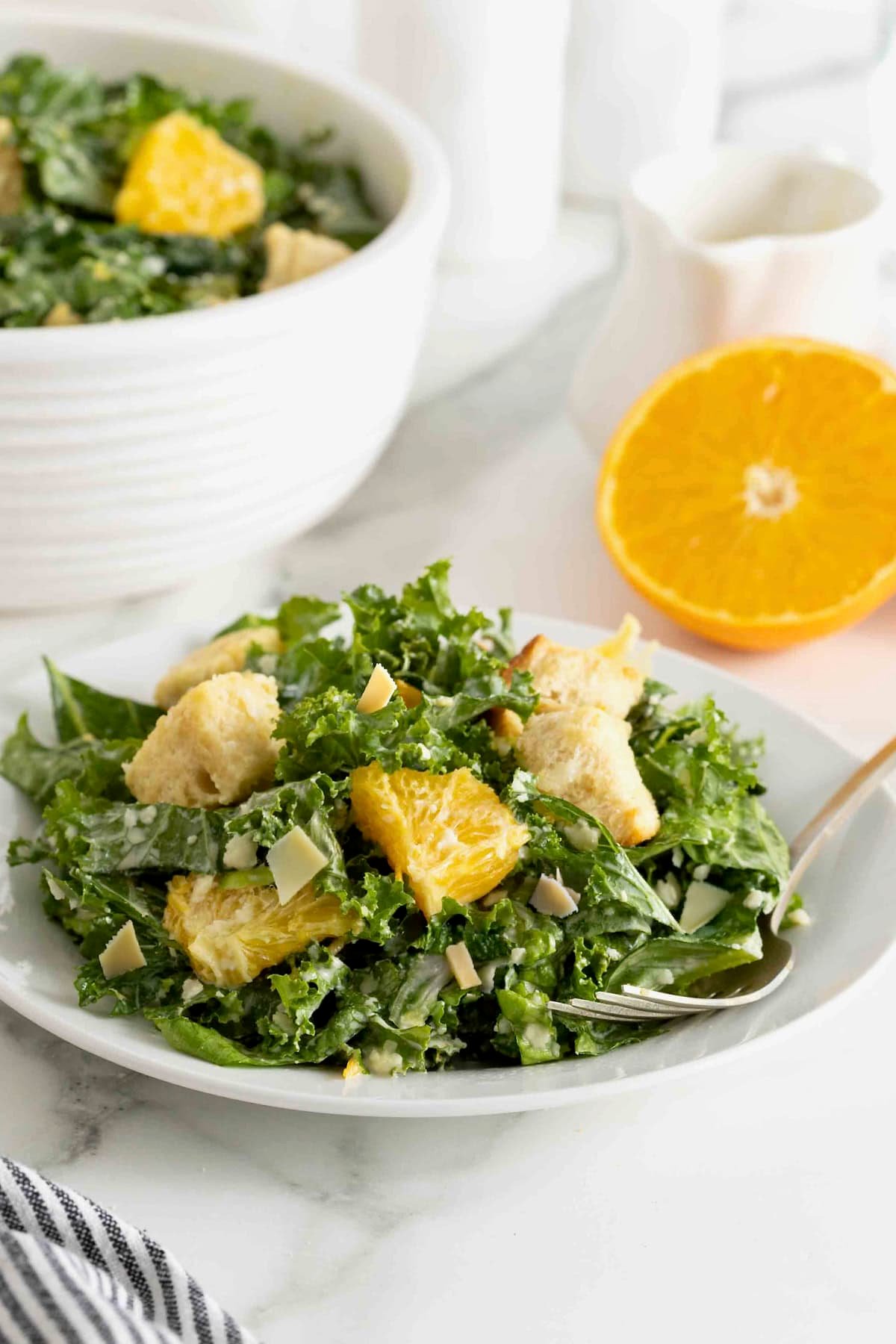 A white salad plate heaped with. citrus caesar salad. There is a fork resting on the plate and a halved orange to the upper right.