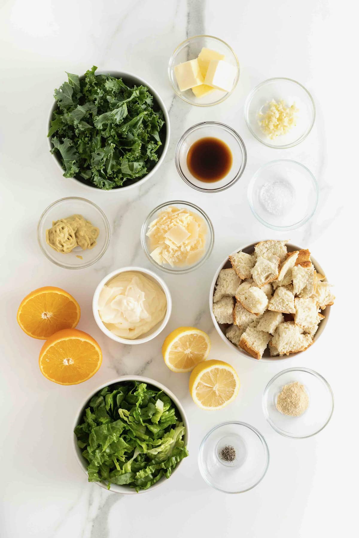 Ingredients to make citrus caesar salad in small glass dishes on a white marble counter.