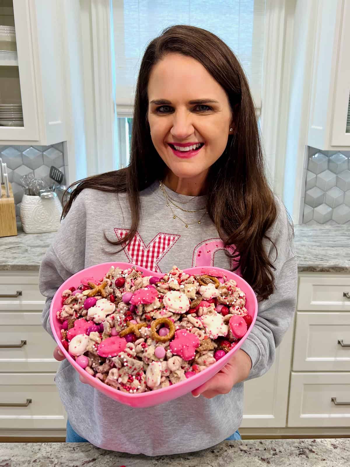 Maegan Brown wearing a light gray sweatshirt with a red x and a pink o on the front. Maegan is holding a pink heart dish of valentine's snack mix.