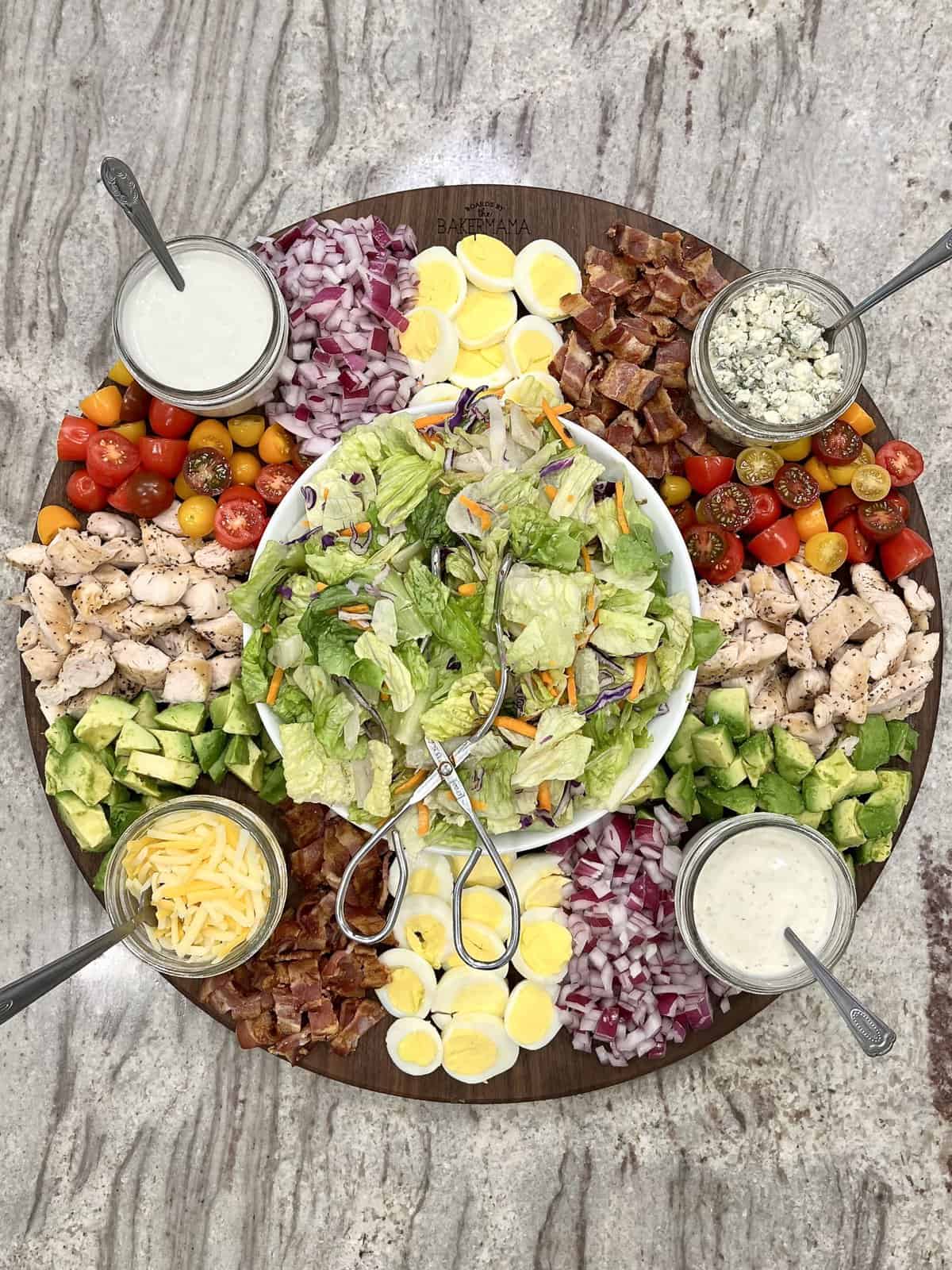 A large round wood board with a bowl of salad in the center surrounded by toppings and salad dressings in small glass jars.