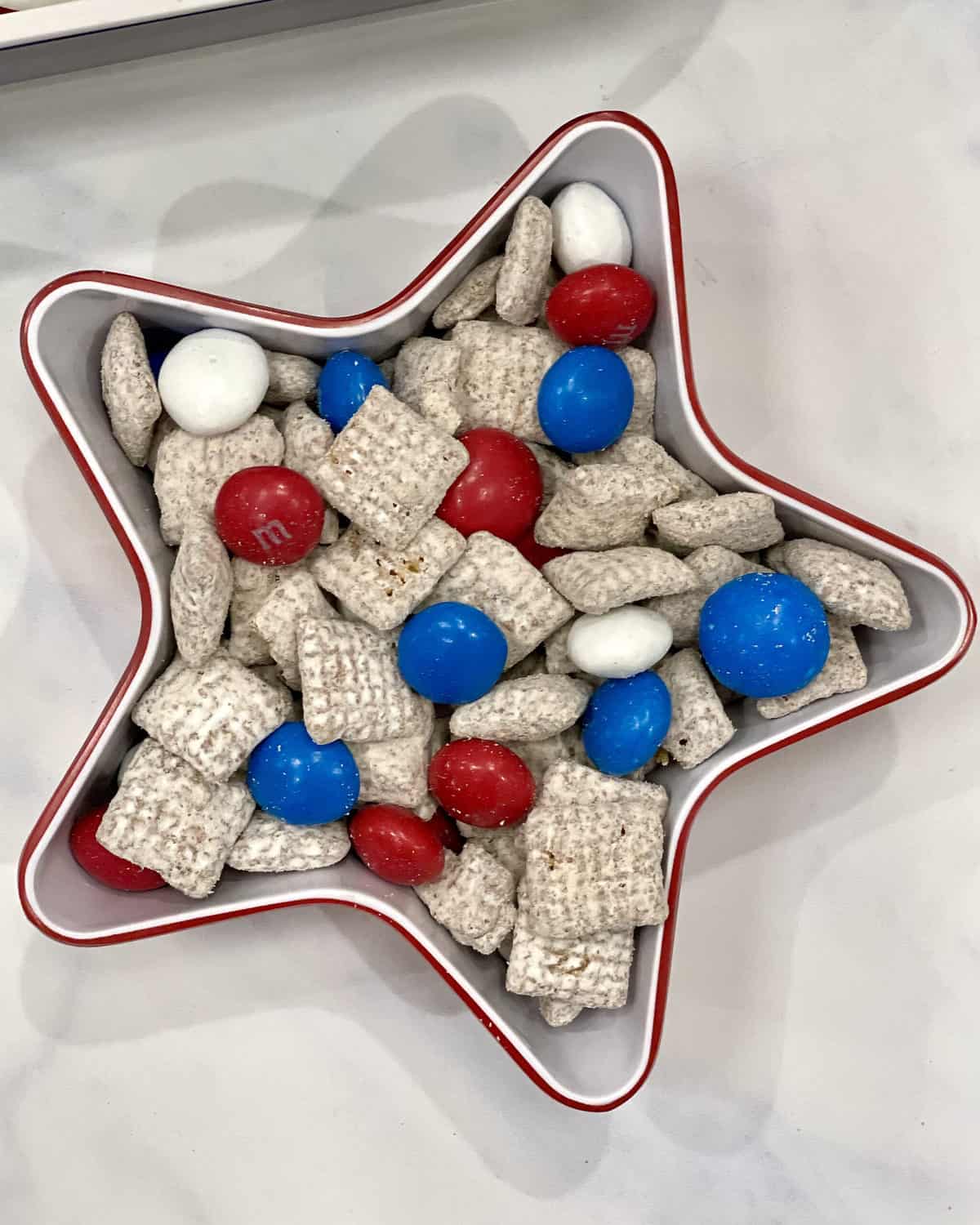 Snack mix made with muddy  buddy buddies, red, white and blue M&Ms in a red and white plastic star-shaped bowl.
