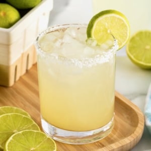 A salt rimmed glass filled with margarita on a rimmed wooden serving tray. There is a lime wedge on the rim of the glass.